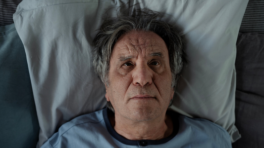 Elderly man lying awake in bed struggling with frequent nighttime urination and sleep disruption.